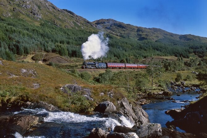 A steam train with maroon coaches in a wooded glen above a rocky river