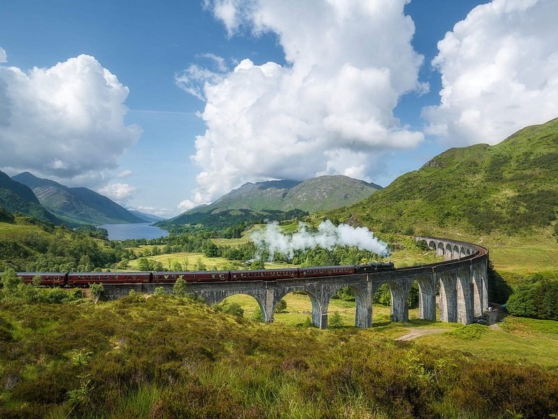 A steam train crossing a curved concrete viaduct, a long loch in the glen beyond